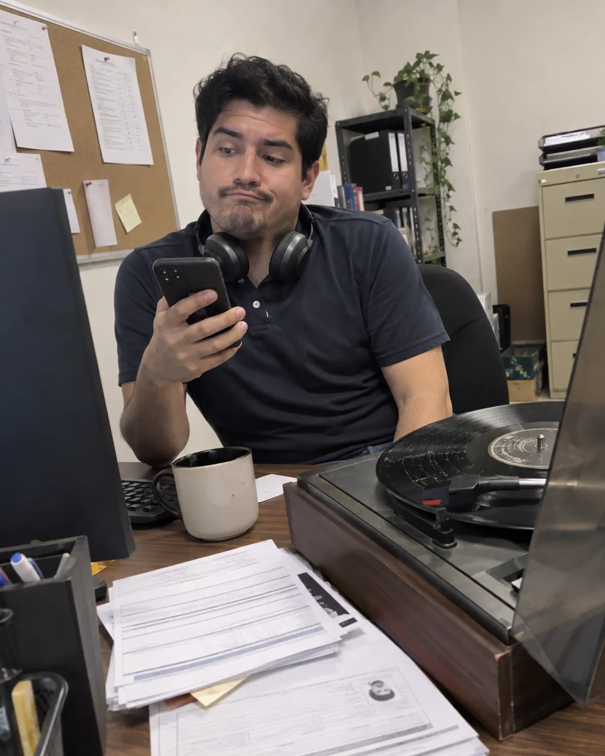 Reclutador en su escritorio con audífonos al cuello y el celular en la mano, con cara de «otra vez lo mismo», junto a un tocadiscos con un disco rayado.